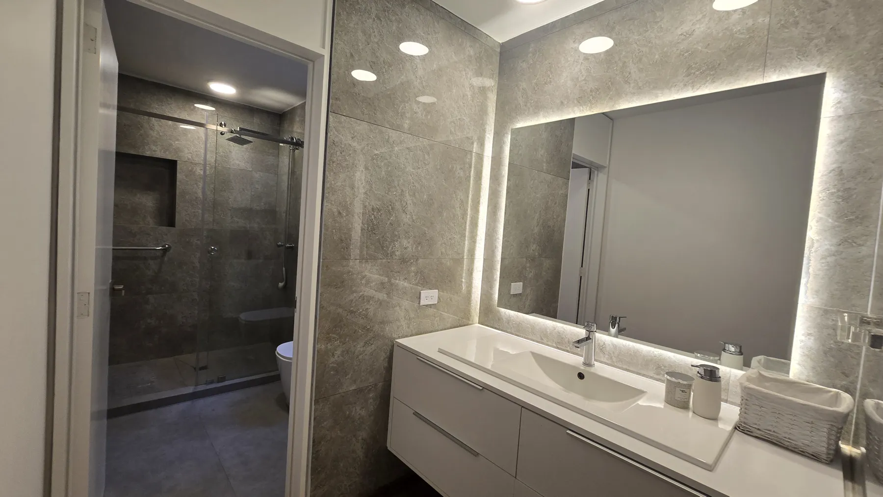 Baño remodelado por Oscar: ducha de vidrio, porcelanato gris y mueble de lavamanos con espejo iluminado